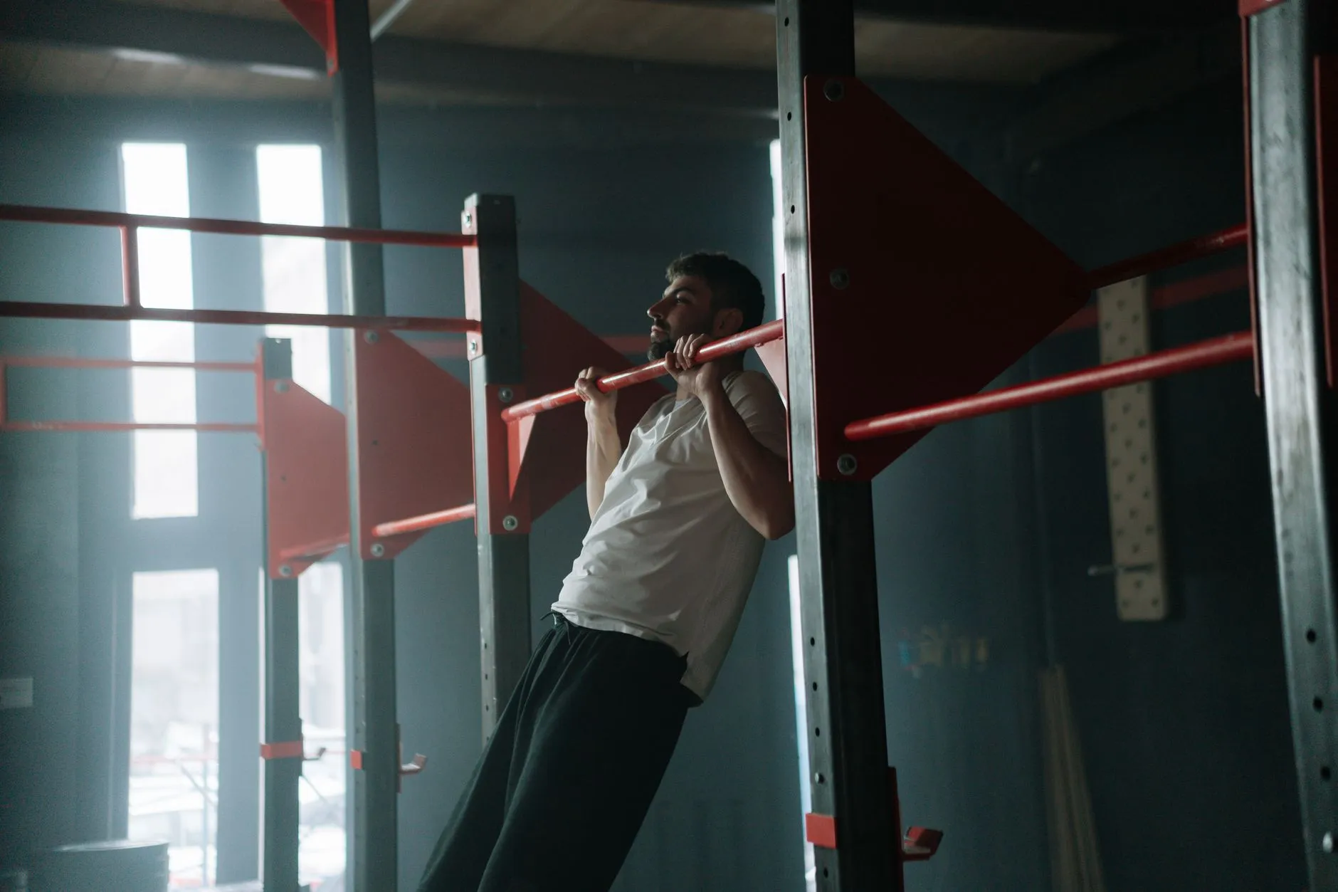 A bearded man pulling himself up on a red pull-up bar in a dim gym, the same movement a doorway pull up bar brings home