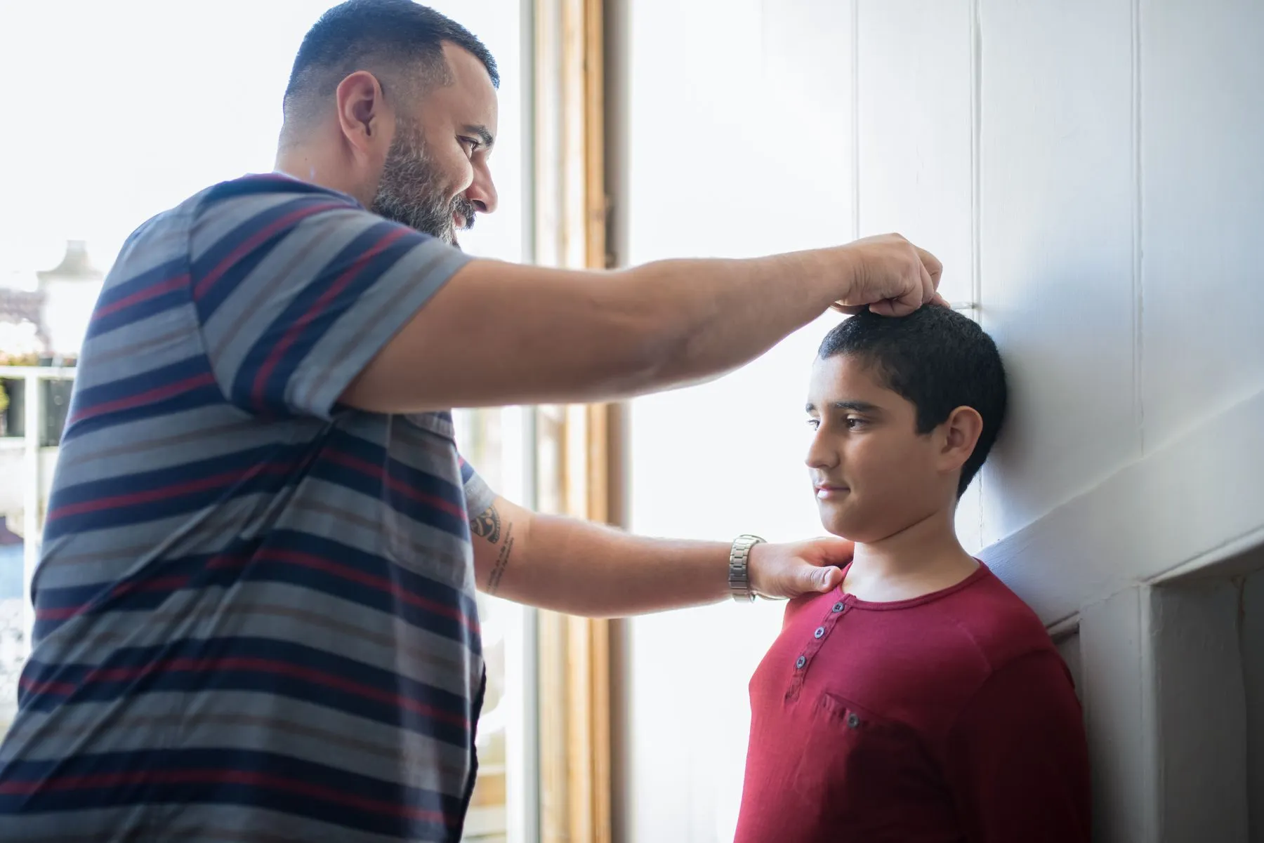 A father marking his teenage son's height against a white wall at home, one hand flat on the boy's head