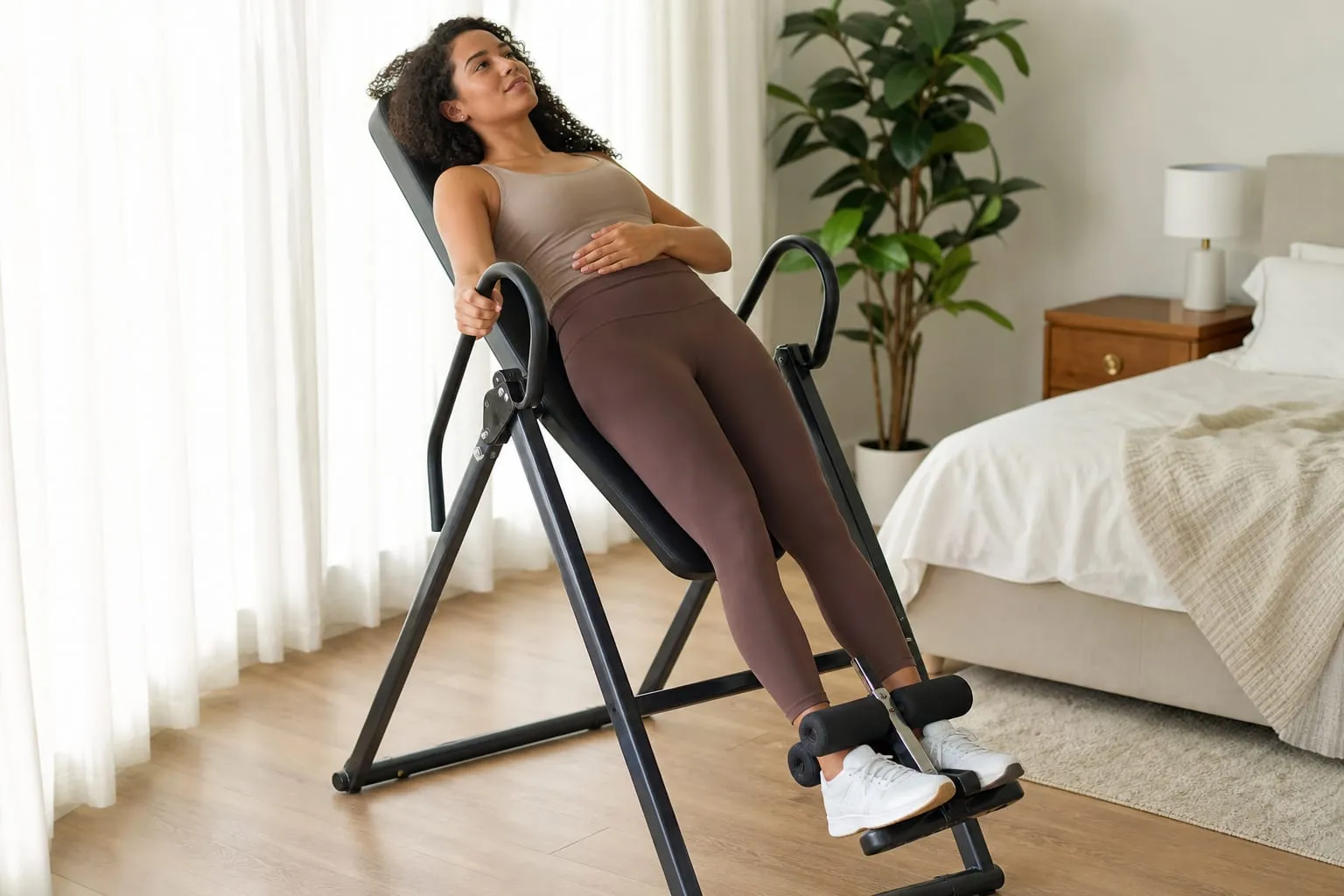 A woman tilted back on an inversion table at a shallow angle in a bright bedroom, one hand on the handle