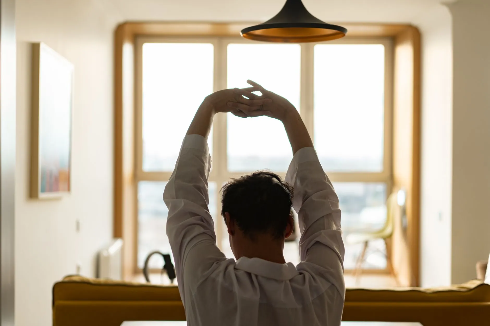 A man seen from behind stretching both arms overhead in a sunlit living room, the kind of home space where a Teeter EP-560 inversion table would stand